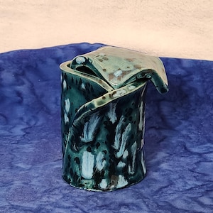 May include: A handcrafted ceramic vase with a unique, folded design. The vase is cylindrical with a folded top, and is glazed in shades of teal and dark blue, creating a marbled effect. The vase is approximately 6 inches tall.