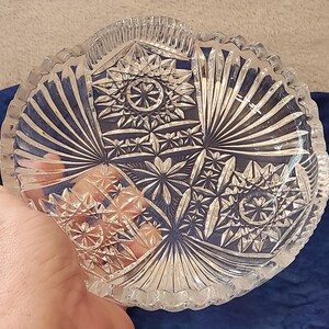 May include: Clear glass bowl with a starburst and geometric design. The bowl has a scalloped edge. The bowl is held by a hand. The bowl is likely vintage and suitable for serving or display.