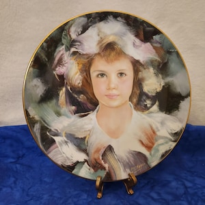 May include: A porcelain plate with a painted image of a young girl with long, flowing hair. The girl is wearing a white dress and has a soft, dreamy expression on her face. The background is a swirling mix of colors, creating a sense of movement and whimsy.