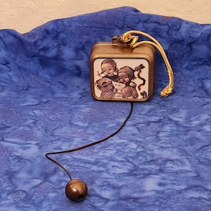May include: A small, square, gold-colored music box with a pull string. The music box has a picture of three children singing on the front. The music box is on a blue fabric background.