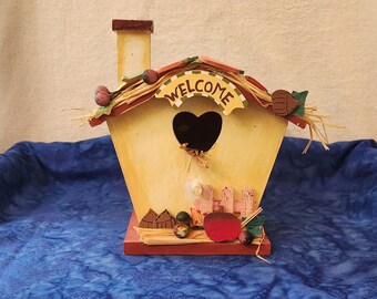 May include: A decorative birdhouse with a heart-shaped opening and the word "WELCOME" on a banner. The birdhouse is painted yellow with a red base and is adorned with faux berries and straw.