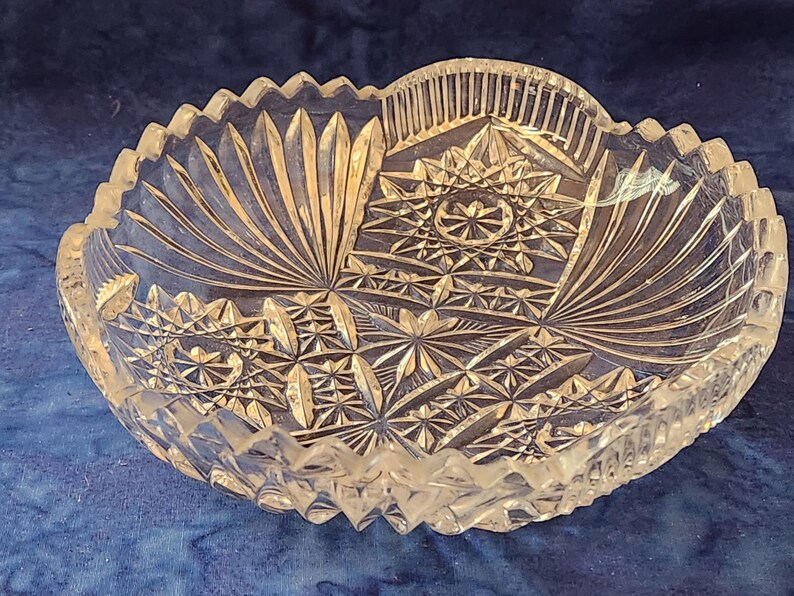 May include: A clear, cut-glass bowl with a scalloped edge. The bowl has intricate geometric and fan-shaped patterns. The design is symmetrical, with a starburst pattern in the centre. A decorative piece for serving or display.