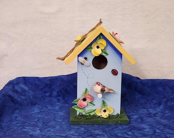 May include: A decorative birdhouse painted light blue with a yellow roof and floral accents. The birdhouse features a round entry hole, a small bird figurine, a ladybug, and a bee. The base is green with faux moss.