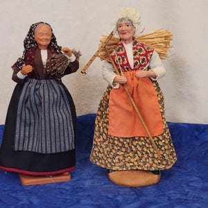 May include: Two vintage folk art dolls depicting elderly women in traditional attire. One doll holds a bundle of twigs, while the other carries a rake and a bundle of straw. The dolls wear aprons, headscarves, and long skirts. The dolls are standing on a blue surface.