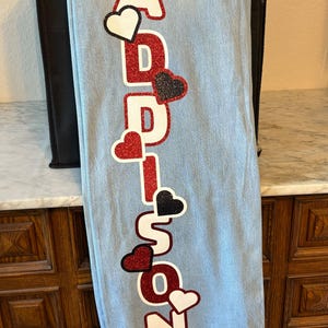 May include: Light blue denim tote bag with the name "ADDISON" vertically displayed in red glitter letters. Black, white, and red heart accents are interspersed throughout the name, adding a decorative touch to the bag.