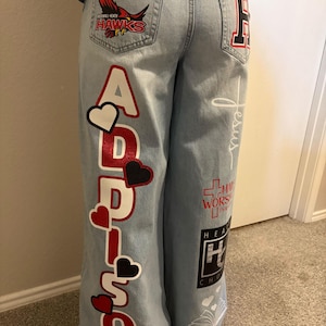 May include: Light blue denim jeans featuring custom graphics including the name Addison in red and white, red and black hearts, a Rockwall-Heath Hawks logo on the back pocket, a large letter H, a cross, the word Jesus, and a book illustration.