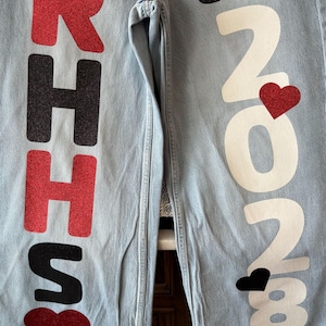 May include: Light blue denim pants with large red glitter letters spelling "RHS" and a red heart. The other leg has the text "c/o 2023" in white with a red glitter heart and a black heart.