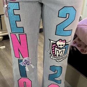 May include: Light wash denim jeans featuring custom glittery lettering and graphics. The right leg displays the word SENIOR in alternating pink and blue glitter. The left leg features the text Class of 26 with the Monster High logo and skull.