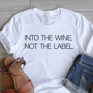 May include: A white t-shirt with the text "INTO THE WINE, NOT THE LABEL." is paired with denim shorts, brown sandals, and sunglasses. The items are arranged on a light wood-grain surface.