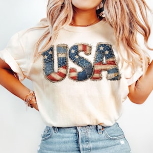 May include: Cream-colored t-shirt with the word "USA" in a patchwork design featuring the American flag. The letters are outlined with a frayed edge, and the shirt is paired with blue denim jeans.