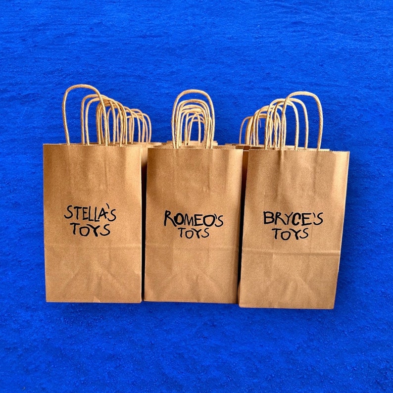 Op de afbeelding: Drie bruine papieren cadeauzakken met handvatten. De zakken zijn gelabeld met de tekst "Stella's Toys", "Romeo's Toys" en "Bryce's Toys".