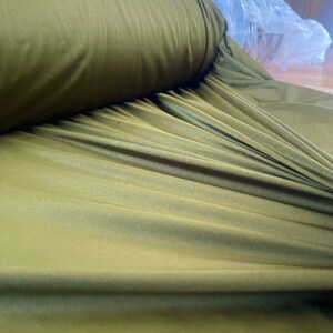 May include: A close-up of a roll of olive green fabric with a smooth, shiny texture. The fabric is folded and draped, showing its soft, flowing quality.