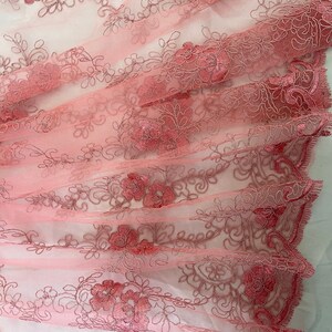 May include: Pink floral lace fabric with a delicate embroidered design. The lace is sheer and has a soft, flowing texture.