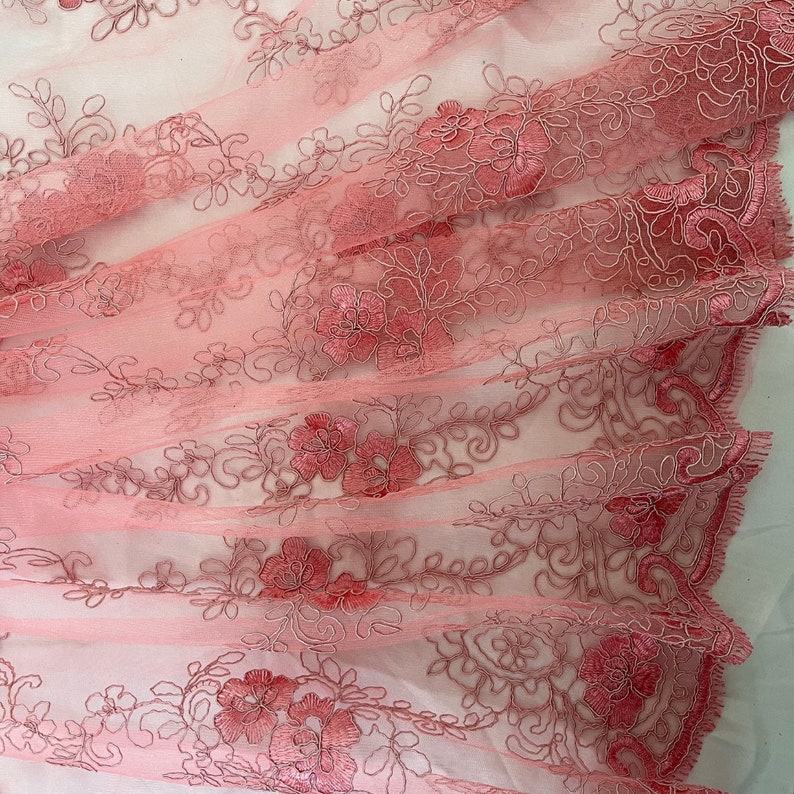 May include: Pink floral lace fabric with a delicate embroidered design. The lace is sheer and has a soft, flowing texture.