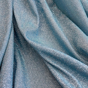 May include: Close-up of a light blue fabric with a shimmery, glittery texture. The fabric is draped and folded, creating soft, flowing lines.