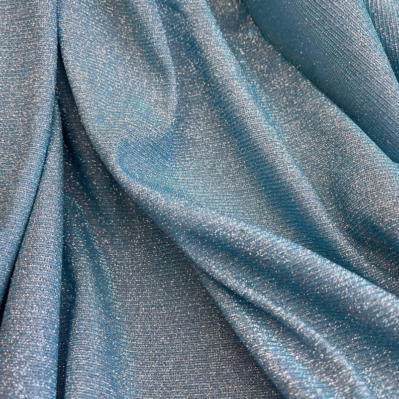 May include: Close-up of a light blue fabric with a shimmery, glittery texture. The fabric is draped and folded, creating soft, flowing lines.