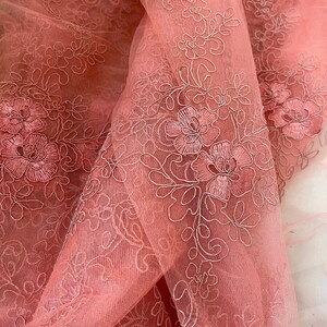 May include: Pink tulle fabric with white embroidered floral designs. The fabric is sheer and delicate, with a soft, romantic feel.
