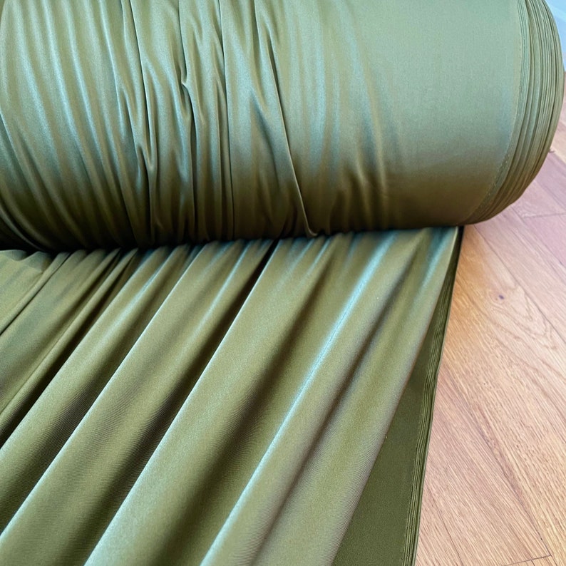 May include: A roll of olive green fabric with a smooth, silky texture. The fabric is folded and draped on a wooden surface.
