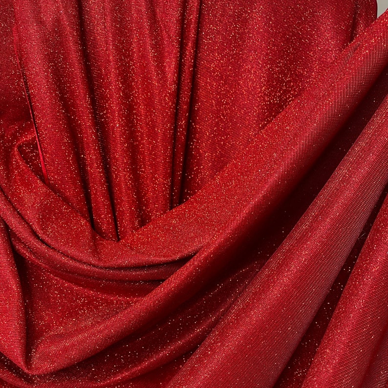 May include: Red sparkly fabric with a fine, shimmery texture. The fabric is draped and folded, showcasing its soft, flowing quality.