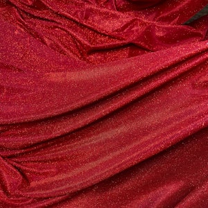May include: Red fabric with a sparkly, glittery texture. The fabric is folded and draped, showing the texture and sheen.