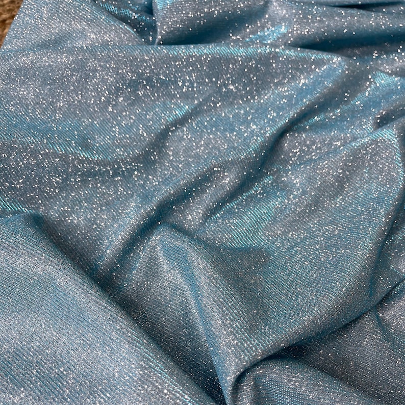 May include: Close-up of a light blue fabric with a sparkly, iridescent finish. The fabric has a subtle ribbed texture.