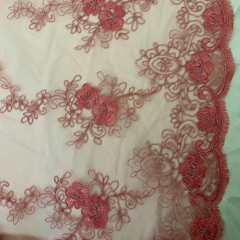 May include: Pink floral lace fabric with a delicate pattern of flowers and leaves. The lace has a scalloped edge.