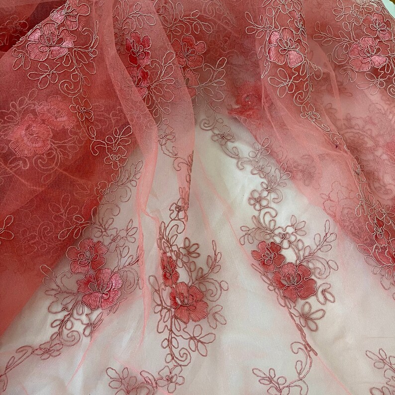 May include: Pink floral lace fabric with a delicate embroidered design. The fabric is sheer and has a soft, flowing texture.