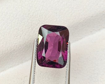 May include: A purple gemstone held by tweezers in a black and white box. The gemstone is a rectangular shape with a smooth, polished surface.
