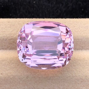 May include: A light pink gemstone with a cushion cut. The gemstone is set on a beige velvet background.
