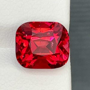 May include: A vibrant, faceted red gemstone with a cushion cut. The gem is a deep, rich red color and appears to be highly polished, reflecting light. The shape is a rounded square, showcasing the intricate internal facets.
