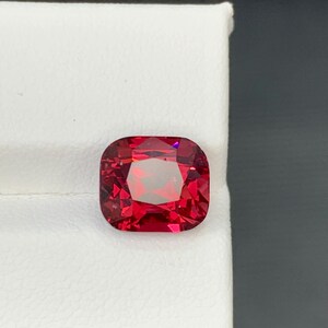 May include: A faceted, cushion-cut gemstone with a vibrant red hue. The gem is set against a white background, showcasing its clarity and brilliance. The square shape with rounded corners is a classic design.