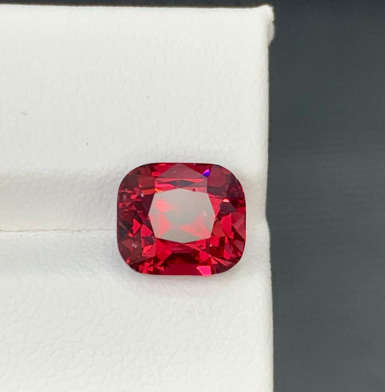 May include: A faceted, cushion-cut gemstone with a vibrant red hue. The gem is set against a white background, showcasing its clarity and brilliance. The square shape with rounded corners is a classic design.