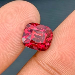 May include: A faceted, cushion-cut gemstone with a vibrant, deep red hue. The gem is held between two fingers, showcasing its brilliance and clarity. The cut of the stone creates multiple facets that reflect light, enhancing its sparkle.