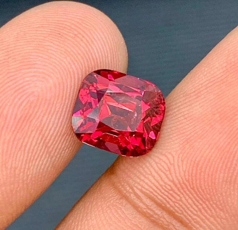 May include: A faceted, cushion-cut gemstone with a vibrant, deep red hue. The gem is held between two fingers, showcasing its brilliance and clarity. The cut of the stone creates multiple facets that reflect light, enhancing its sparkle.