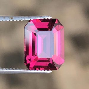 May include: A vibrant, deep pink gemstone cut in a rectangular emerald shape with faceted surfaces. The stone is held by a pair of silver-toned metal tweezers, showcasing its translucent clarity and sharp, geometric edges against a neutral background.