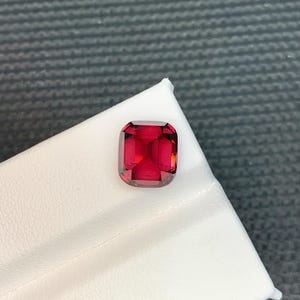 May include: A vibrant, cushion-cut, red gemstone. The gem is set against a white surface, with a dark gray background. The gemstone's facets reflect light, showcasing its clarity and rich color. This gemstone is suitable for jewelry making.