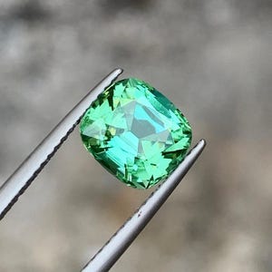 May include: A cushion-cut green gemstone with a slight yellow hue, held in tweezers.