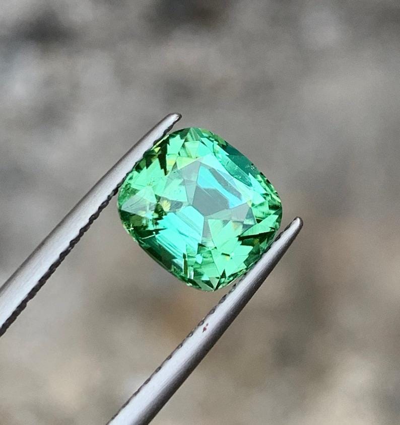 May include: A cushion-cut green gemstone with a slight yellow hue, held in tweezers.