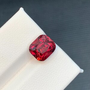 May include: A faceted, cushion-cut gemstone in a deep red hue. The gem is displayed on a white surface, with a dark gray background. The gemstone's cut reveals multiple facets, reflecting light.
