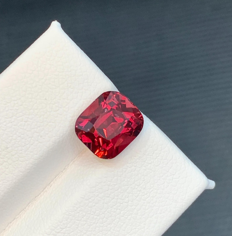 May include: A faceted, cushion-cut gemstone in a deep red hue. The gem is displayed on a white surface, with a dark gray background. The gemstone's cut reveals multiple facets, reflecting light.