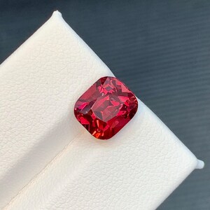 May include: A vibrant, cushion-cut gemstone with a deep red hue. The gem is set against a white surface, showcasing its facets and brilliance. The square shape with rounded corners is a classic design.