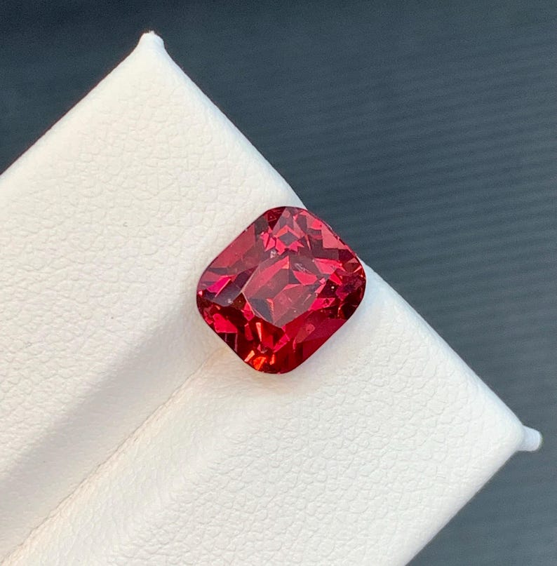 May include: A vibrant, cushion-cut gemstone with a deep red hue. The gem is set against a white surface, showcasing its facets and brilliance. The square shape with rounded corners is a classic design.