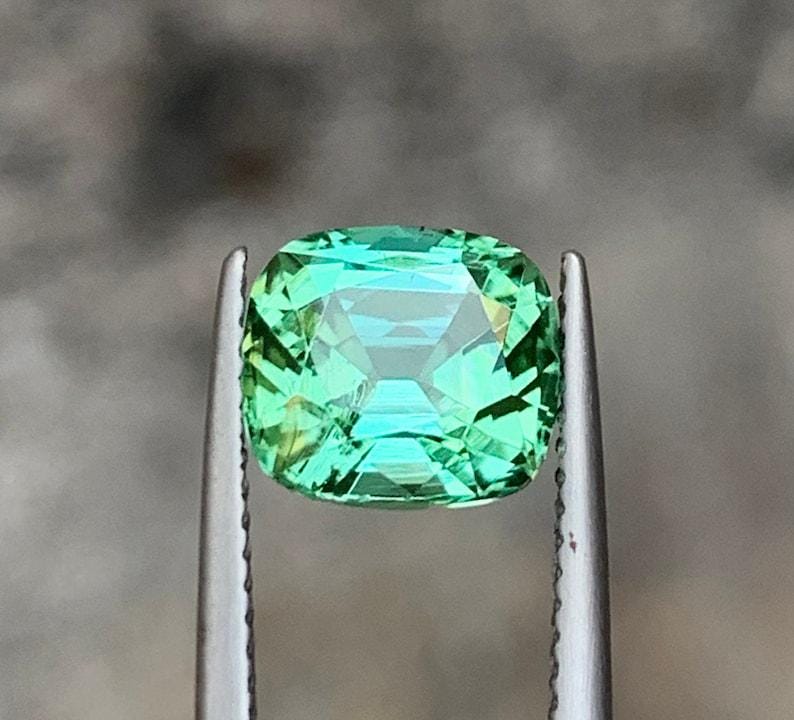 May include: A green gemstone with a cushion cut. The gemstone is held in place by a pair of tweezers.