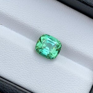 May include: A cushion-cut green gemstone with a slight yellow hue, set in a white ring box.