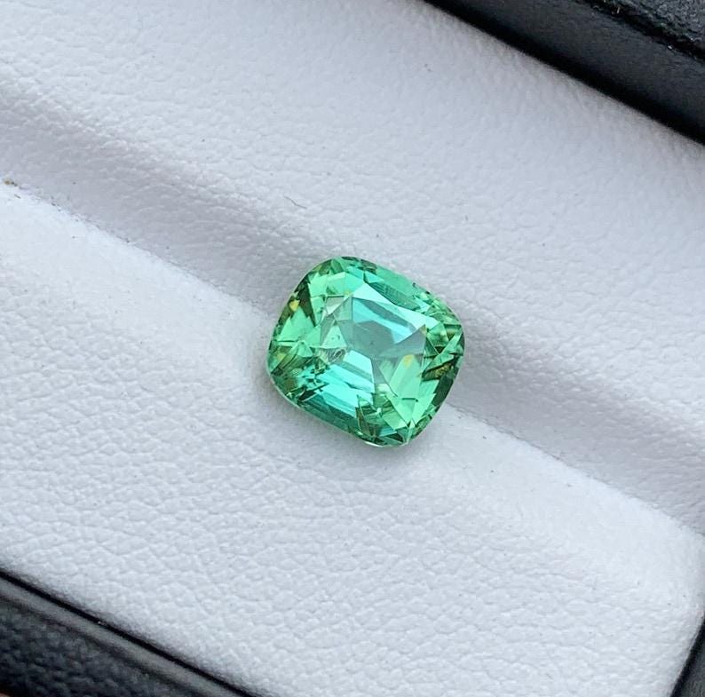 May include: A cushion-cut green gemstone with a slight yellow hue, set in a white ring box.