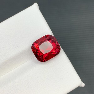 May include: A vibrant, cushion-cut red gemstone with a brilliant, faceted surface. The gem is displayed on a white surface, with a dark gray background. The gemstone's color is a deep, saturated red, reflecting light.