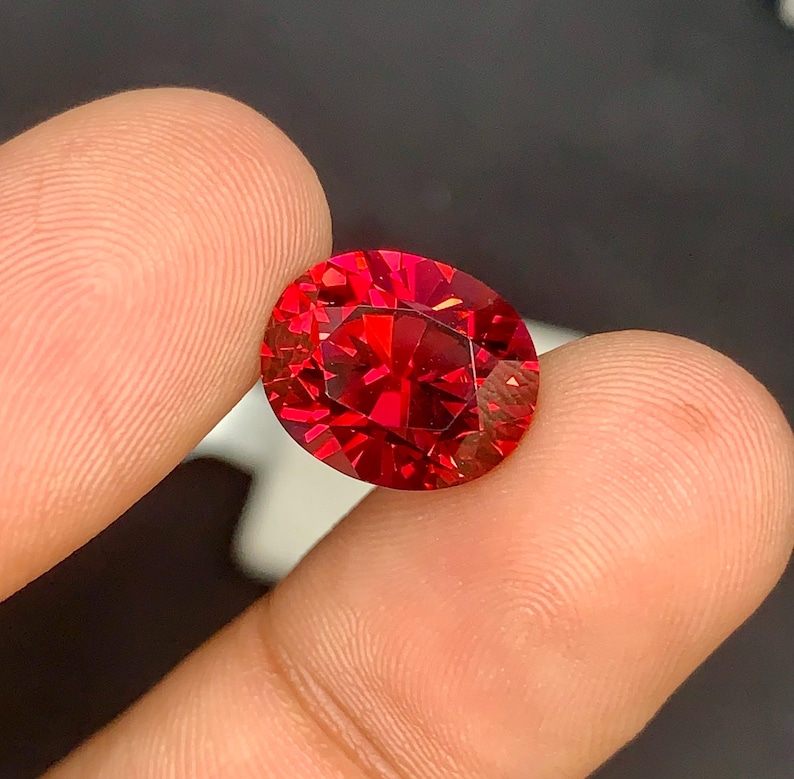 May include: A faceted, oval-cut gemstone with a vibrant red hue. The gem is held between two fingers, showcasing its brilliance and intricate internal structure. The gem's facets reflect light, creating a sparkling effect. The background is dark, emphasizing the gem's color and clarity.