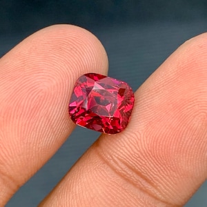 May include: A faceted, cushion-cut gemstone in a vibrant, deep red hue. The gem is held between two fingers, showcasing its polished surfaces and internal reflections. The stone's shape is square with rounded corners, and it appears to be a high-quality jewel.