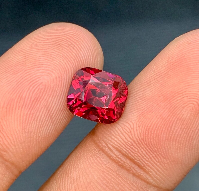 May include: A faceted, cushion-cut gemstone in a vibrant, deep red hue. The gem is held between two fingers, showcasing its polished surfaces and internal reflections. The stone's shape is square with rounded corners, and it appears to be a high-quality jewel.