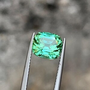 May include: A green gemstone with a cushion cut, held by tweezers. The gemstone has a slight blue hue.